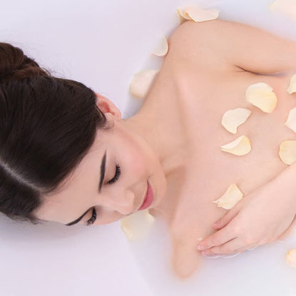 Eine Frau mit dunklen Haaren entspannt in einem Milchbad mit hellen Rosenblättern auf der Haut.