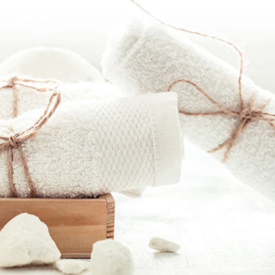 Weißes Handtuchset mit dekorativer Kordel auf einem Holztablett, daneben weiße Naturseife und Bimssteine.
