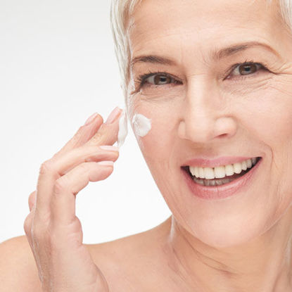 Eine lächelnde ältere Frau mit kurzen grauen Haaren trägt sich sanft Gesichtscreme auf die Wange auf. Sie blickt direkt in die Kamera und vermittelt ein Gefühl von Zufriedenheit und Pflege.