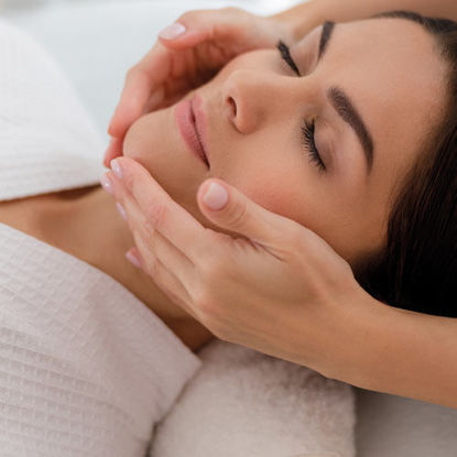 Eine entspannte Frau mit geschlossen Augen erhält eine sanfte Gesichtsmassage in wohltuender Umgebung.