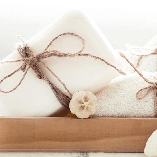 Ein dekoratives Arrangement aus weißer Seife, gebunden mit rustikalem Juteband, daneben ein weiches, gefaltetes Handtuch und eine kleine getrocknete Blume. Alles präsentiert auf einem Holztablett in heller, natürlicher Umgebung.