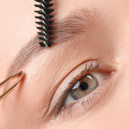 Nahaufnahme eines Auges mit einer Pinzette, die ein Augenbrauenhaar zupft, und einer Augenbrauenbürste, die die Braue formt.