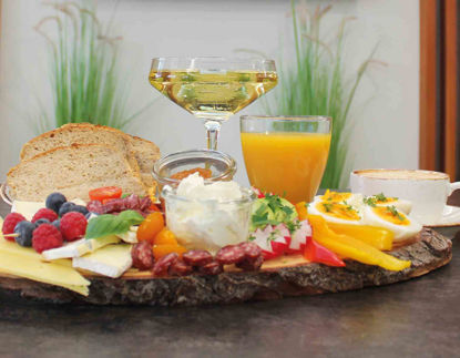 Ein liebevoll angerichtetes Frühstücksbrett mit Aufschnitt, Käse, frischem Gemüse, Obst und Dips steht auf einem rustikalen Holztablett. Daneben ein Glas Orangensaft, ein Glas Sekt und ein Korb mit Körnerbrot. Im Hintergrund ein stilvoll eingerichteter Raum mit Holzverkleidung und dekorativen Schneidebrettern an der Wand.