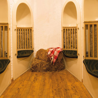 Ein gemütlich eingerichteter Ruheraum mit Holzfußboden, halbrunden Nischen mit gepolsterten Sitzflächen und Heuballen, auf denen eine rot-karierte Decke liegt. Die Atmosphäre ist warm und einladend.
