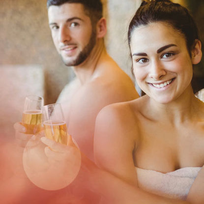 Ein junges Paar sitzt in einer Wellnessumgebung, beide tragen weiße Handtücher. Sie halten Sektgläser in der Hand und lächeln in die Kamera. Die Stimmung ist entspannt und freundlich, im Hintergrund ist eine warme, natursteinartige Wand zu erkennen.