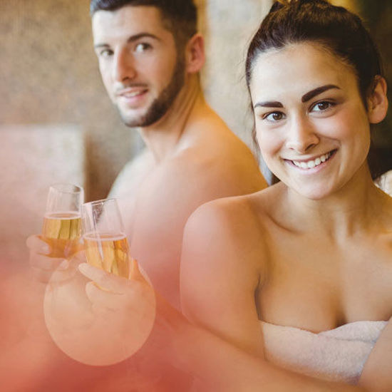 Ein junges Paar sitzt in einer Wellnessumgebung, beide tragen weiße Handtücher. Sie halten Sektgläser in der Hand und lächeln in die Kamera. Die Stimmung ist entspannt und freundlich, im Hintergrund ist eine warme, natursteinartige Wand zu erkennen.