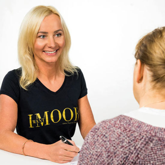 Zwei Frauen im Gespräch, eine davon trägt ein schwarzes T-Shirt mit der Aufschrift „I WANT MOOR“ und hält einen Stift in der Hand.