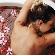 Eine Frau entspannt in einer runden Badewanne mit warmem Wasser und schwimmenden Blüten. Sie lehnt sich mit geschlossenen Augen und erhobenen Armen an den Wannenrand, umgeben von Wellnessprodukten. Die Atmosphäre wirkt ruhig und erholsam.