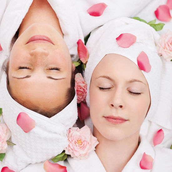 Zwei Frauen in weißen Bademänteln und Handtuchturbanen liegen entspannt mit geschlossenen Augen, umgeben von Rosenblättern und Blüten.