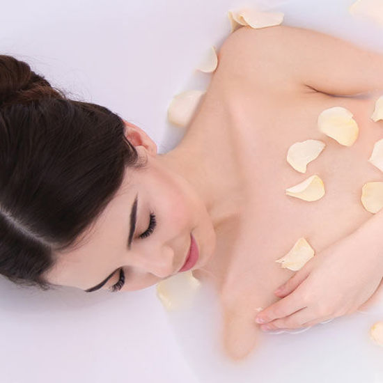 Eine Frau mit dunklen Haaren entspannt in einem Milchbad mit hellen Rosenblättern auf der Haut.