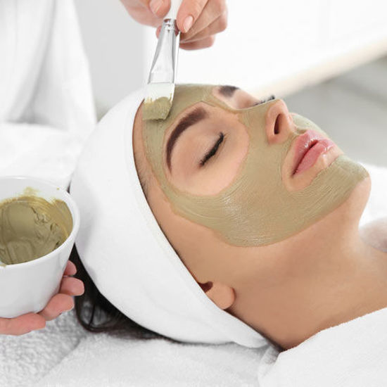 Eine Frau liegt entspannt auf einer Liege mit geschlossen Augen, während ihr mit einem Pinsel eine grüne Gesichtsmaske aufgetragen wird. Sie trägt ein weißes Haarband und ist in ein weißes Handtuch gehüllt.
