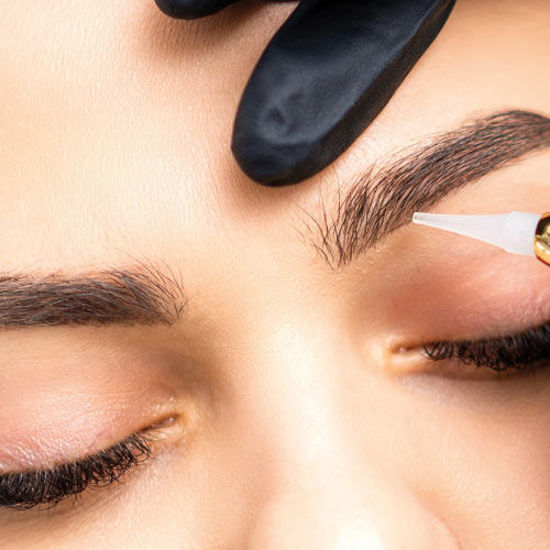 Eine Frau erhält eine Augenbrauenbehandlung. Eine Hand mit schwarzem Handschuh hält ihre Stirn, während mit einem feinen Stift Produkt auf die Augenbraue aufgetragen wird.