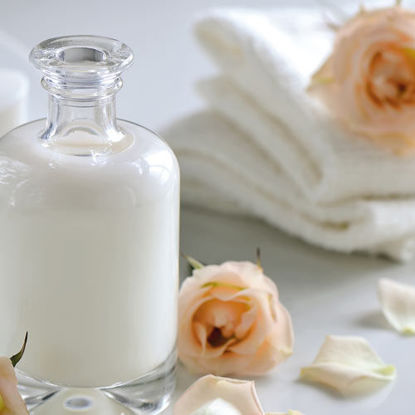Eine elegante Glasflasche mit weißer Lotion steht auf einem hellen Untergrund. Umgeben ist sie von zarten Rosenblüten und weißen Handtüchern, was eine ruhige, pflegende Spa-Atmosphäre vermittelt.