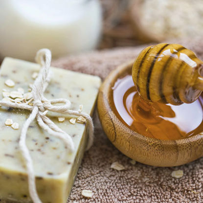 Ein Stück handgemachte Seife mit Haferflocken liegt auf einem Handtuch neben einer Holzschale mit flüssigem Honig und einem Honiglöffel.
