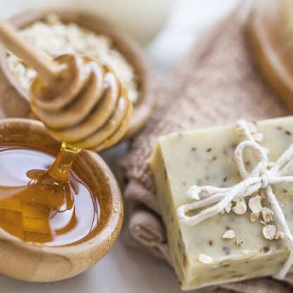 Naturseife mit Haferflocken dekoriert, daneben eine Schale mit flüssigem Honig und ein Honiglöffel arrangiert auf einem weichen Handtuch.