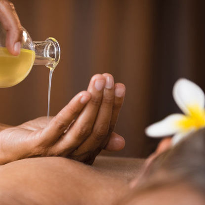 Warmes Massageöl wird in die Hände einer Person gegossen, im Vordergrund liegt eine entspannte Person mit einer Blume im Haar.