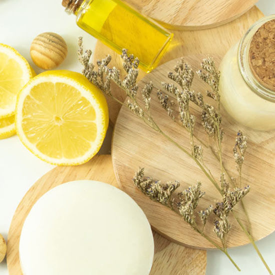Arrangement aus Naturkosmetik-Zutaten: eine aufgeschnittene Zitrone, Massagekugeln aus Holz, ein Glas mit gelbem Öl, getrocknete Lavendelzweige und weitere Naturprodukte auf runden Holzunterlagen.