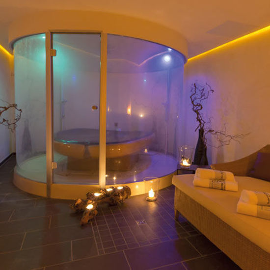 Ein stilvoll beleuchteter Wellnessraum mit einer modernen Membranliege in einer gläsernen Kabine. Umgeben von dekorativen Elementen wie Ästen und Kerzenlicht entsteht eine warme und entspannte Atmosphäre. Daneben liegt ein Handtuch auf einer bequemen Duoliege.