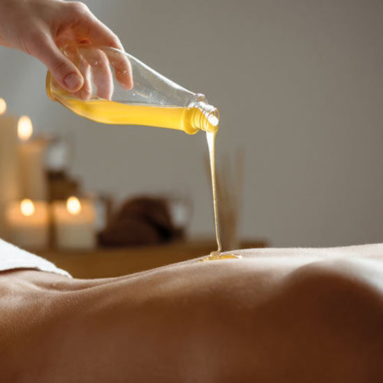 Eine Hand gießt warmes Massageöl aus einer Glasflasche auf den Rücken einer liegenden Person. Im Hintergrund sorgen brennende Kerzen für eine entspannte und wohltuende Atmosphäre.