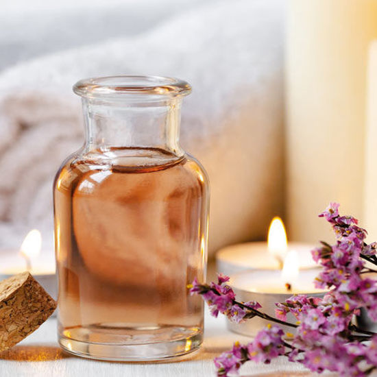 Ein Glasfläschchen mit rosafarbenem Öl steht auf einem Tisch, umgeben von brennenden Teelichtern, einem Handtuch, einem Korken und violetten Blüten.