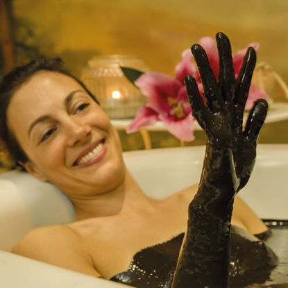 Frau entspannt in einem Moorbad, hebt lächelnd ihre mit Moor bedeckte Hand, im Hintergrund brennende Kerzen und rosa Lilien.
