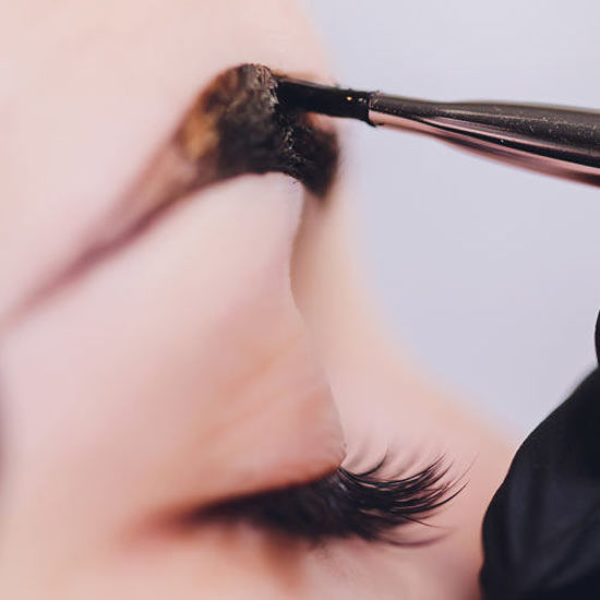 Nahaufnahme eines geschlossenen Auges mit langen Wimpern, während mit einem Pinsel dunkle Farbe auf die Augenbraue aufgetragen wird.