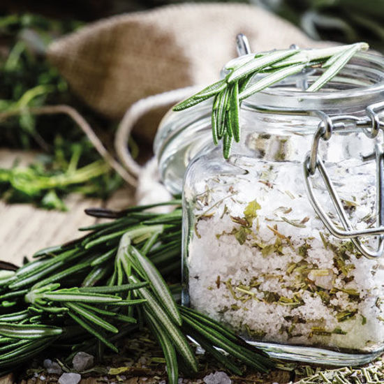 Ein Glas mit grobem Meersalz und getrockneten Kräutern, verziert mit frischem Rosmarin. Im Hintergrund liegen weitere Kräuter und ein Jutesäckchen, was eine natürliche und aromatische Atmosphäre vermittelt.