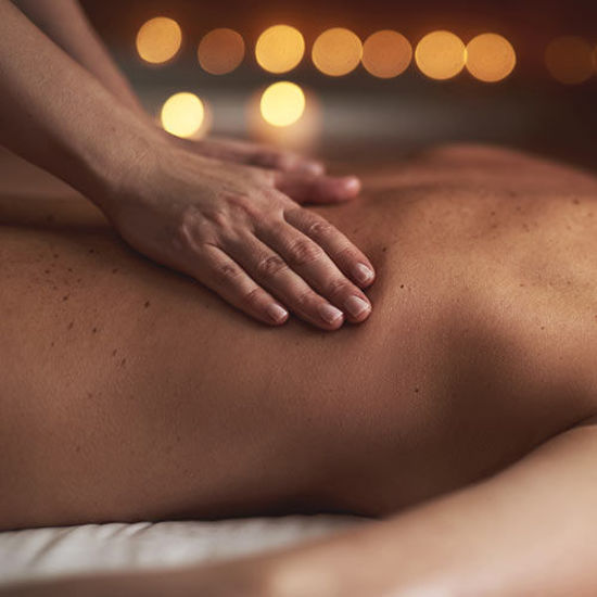 Eine entspannende Rückenmassage bei gedämpftem Licht. Die Hände der Masseurin liegen sanft auf dem entblößten Rücken einer liegenden Person. Im Hintergrund sorgen warme, unscharfe Lichtpunkte für eine beruhigende Atmosphäre.