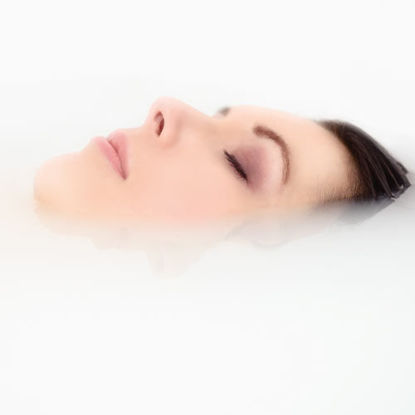Frau mit geschlossenen Augen entspannt im Liegen, ihr Gesicht ragt ruhig aus der milchig-weißen Wasseroberfläche.