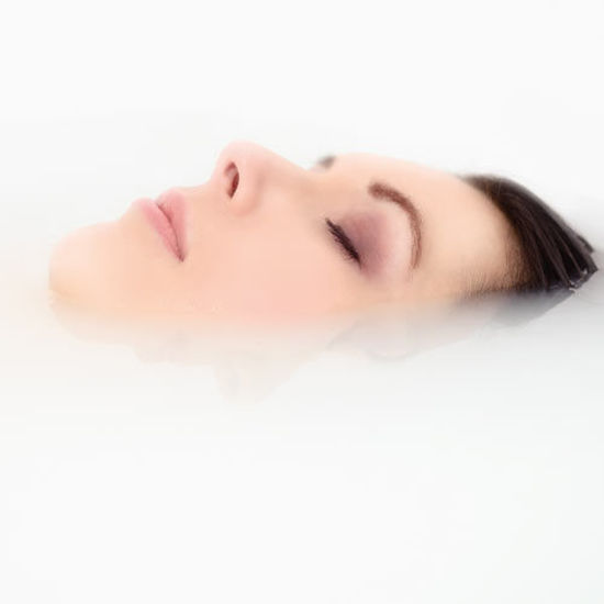 Frau mit geschlossenen Augen entspannt im Liegen, ihr Gesicht ragt ruhig aus der milchig-weißen Wasseroberfläche.