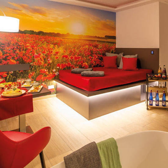 Ausschnitt aus dem Privat Spa. Ein Wasserbett mit roten Dekokissen und einer grauen Kuscheldecke. Das Bett wird von unten mit einem Lichtschlauch warm gelb beleuchtet. Auf der Hintergrundwand ist ein Sonnenuntergang zu sehen und ein Kornblumenfeld in Rot abgebildet. Zwei rote Sesselartige Stühle mit einem gedeckten Tisch.