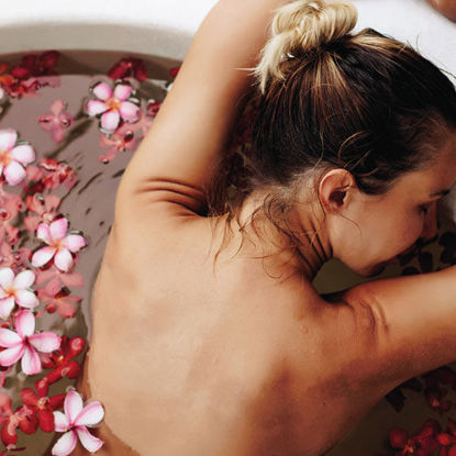 Eine Frau entspannt in einer runden Badewanne mit warmem Wasser und schwimmenden Blüten. Sie lehnt sich mit geschlossenen Augen und erhobenen Armen an den Wannenrand, umgeben von Wellnessprodukten. Die Atmosphäre wirkt ruhig und erholsam.