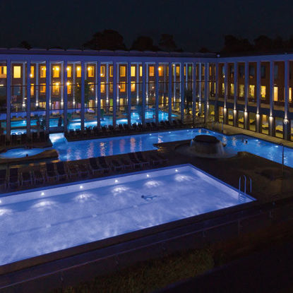 Süßwassersportbecken im Außenbereich der SaarowTherme bei Nacht. Blick auf das beleuchtete Gebäude mit warmem Licht aus den Fenstern, lila gefärbtes Wasser durch Unterwasserscheinwerfer und dunkler Nachthimmel im Hintergrund.