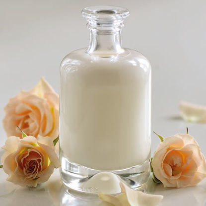 Eine elegante Glasflasche mit weißer Lotion steht auf einem hellen Untergrund, umgeben von zartrosa Rosen und Rosenblättern.
