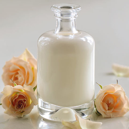 Eine elegante Glasflasche mit weißer Lotion steht auf einem hellen Untergrund, umgeben von zartrosa Rosen und Rosenblättern.