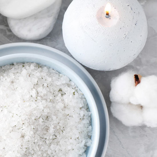 Eine weiße Kugelkerze brennt auf einem hellen Untergrund. Daneben stehen eine Schale mit grobem Badesalz und eine weiche Baumwollblüte.