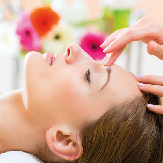 Eine Frau liegt entspannt mit geschlossenen Augen auf einer Wellnessliege, während eine Hand sanft ihre Stirn massiert. Im Hintergrund sorgen bunte Blumen für eine ruhige und angenehme Atmosphäre.