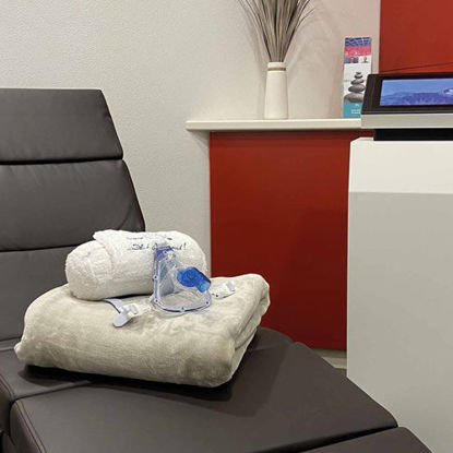 Eine moderne Liege im Hypoxieraum, auf der ein gefaltetes Handtuch, ein Bademantel und eine blaue Atemmaske liegen. Im Hintergrund sind eine rote Wand, ein weißes Regal mit Dekoration und ein Informationsflyer zu sehen.