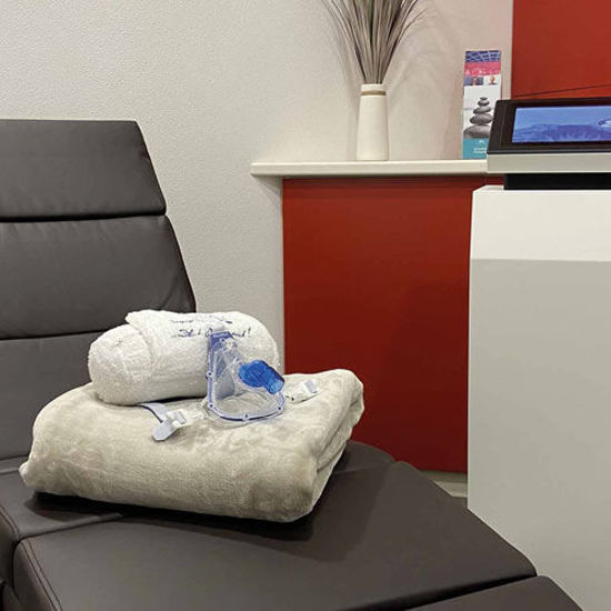 Eine moderne Liege im Hypoxieraum, auf der ein gefaltetes Handtuch, ein Bademantel und eine blaue Atemmaske liegen. Im Hintergrund sind eine rote Wand, ein weißes Regal mit Dekoration und ein Informationsflyer zu sehen.