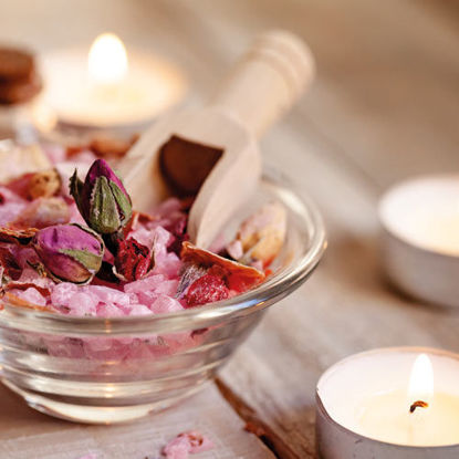 Eine Glasschale mit rosa Badesalz, Rosenknospen und einer kleinen Holzschaufel, umgeben von brennenden Teelichtern auf einem Holztisch.