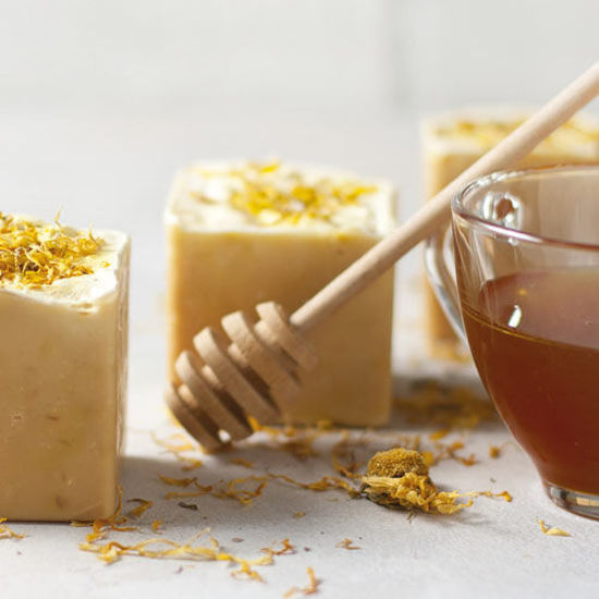 Handgemachte Seifenstücke mit getrockneten Blüten, daneben eine Tasse mit Honig und ein Holz-Honiglöffel.