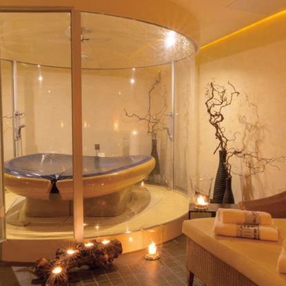 Ein stilvoll beleuchteter Wellnessraum mit einer modernen Wasserliege in einer gläsernen Kabine. Umgeben von dekorativen Elementen wie Ästen und Kerzenlicht entsteht eine warme und entspannte Atmosphäre. Daneben liegt ein Handtuch auf einer bequemen Liege.