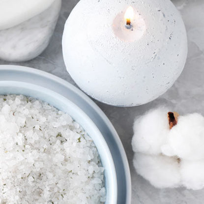 Eine weiße Kugelkerze brennt auf einem hellen Untergrund. Daneben stehen eine Schale mit grobem Badesalz und eine weiche Baumwollblüte.