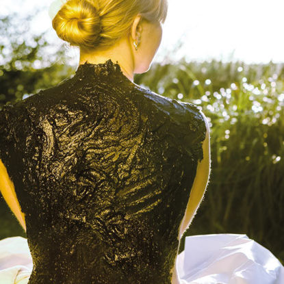 Eine Frau mit blonden Haaren in einem Dutt sitzt im Freien mit dem Rücken zur Kamera, ihr Rücken ist vollständig mit einer schwarzen Moorpackung bedeckt.