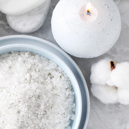Eine weiße Kugelkerze brennt auf einem hellen Untergrund. Daneben stehen eine Schale mit grobem Badesalz und eine weiche Baumwollblüte.