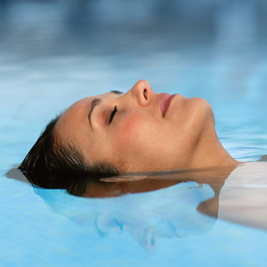 Eine Frau liegt entspannt auf dem Rücken im Wasser eines Pools und hat die Augen geschlossen. Ihr Gesicht ist teilweise unter Wasser, die Wasseroberfläche ist ruhig und spiegelt ihr Profil. Die Atmosphäre wirkt friedlich und erholsam.