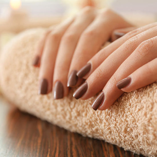 Nahaufnahme gepflegter Frauenhände mit braun lackierten Fingernägeln, die entspannt auf einem beigen Frotteehandtuch ruhen; im Hintergrund sorgen weiche Lichtreflexe für eine warme, entspannende Atmosphäre.