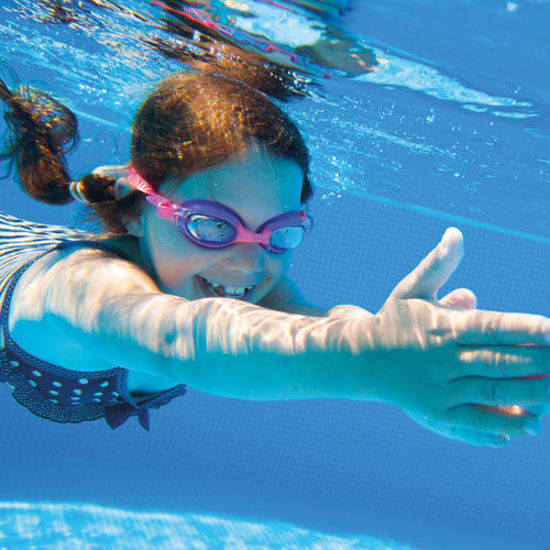 Lachendes Mädchen mit rosa Schwimmbrille taucht unter Wasser in einem Pool und streckt die Arme nach vorne.