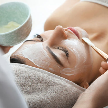 Eine Frau liegt entspannt mit geschlossenen Augen auf einer Liege, während ihr eine weiße Gesichtsmaske mit einem Pinsel aufgetragen wird. Sie ist in ein Handtuch eingehüllt, und eine Schale mit der Maske wird danebengehalten. Die Szene vermittelt eine ruhige Wellness-Atmosphäre.