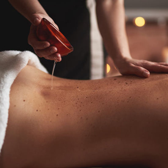 Eine entspannende Aromaölrückenmassage bei Kerzenbeleuchtung. Eine Hand der Masseurin liegt auf dem Rücken einer liegenden Frau. Die andere Hand hält eine Schale mit warmem Aromaöl. Sanft läuft über dem Rücken der liegenden Person das warme Aromaöl. Im Hintergrund sorgen warme, unscharfe Kerzen-Lichtpunkte für eine beruhigende Atmosphäre.